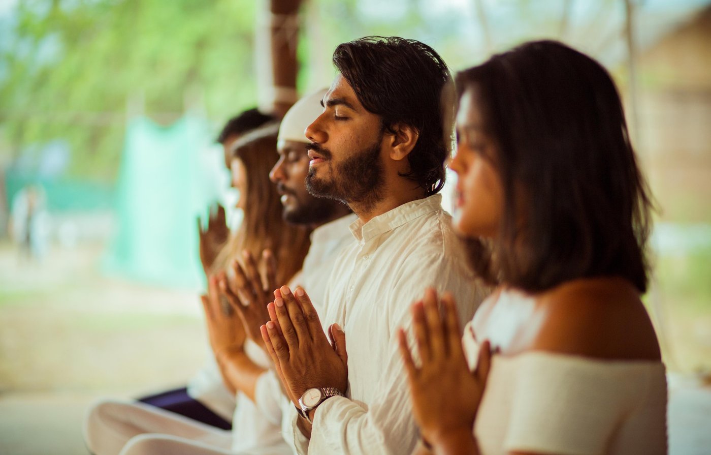 Teachers from the Kashish Yoga family seated in a row with folded hands, eyes closed, in quiet prayer